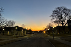 Eine Straße mit Häusern und Bäumen an beiden Seiten und die Sonne geht auf und leuchtet hell orange.