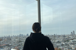 Blick über Nagoya von der Nanzan Universität Katharina, wie sie vor einem großen Eckfenster steht und über Nagoya schaut.