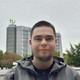 A young man looks into the camera. His head and shoulders are shown. In the background you can see the TU Dortmund's math tower with the TU logo on the roof.