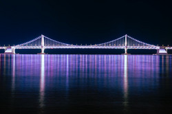 Aussicht auf eine lila-blau beleuchtete Brücke über dem Wasser bei Nacht. Die Lichter spiegeln sich in der Wasseroberfläche. 