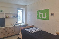 Wohnheimzimmer Hier sieht man das Wohnheimzimmer des Studenten. Man sieht eine Kommode, ein Einzelbett und die TU Flagge über dem Bett.