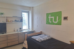 Hier sieht man das Wohnheimzimmer des Studenten. Man sieht eine Kommode, ein Einzelbett und die TU Flagge über dem Bett. 