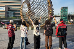 Hier sieht man 5 Menschen mit Baseball Trikots von hinten, die vor einem großen Ball aus Metall stehen.