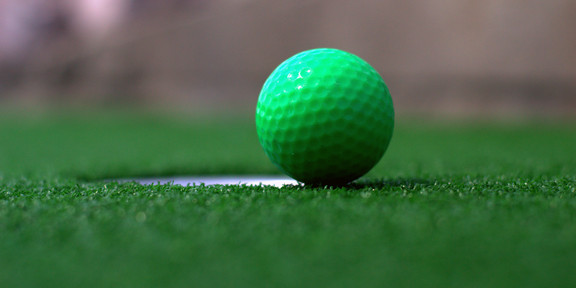 A green miniature golf ball near a hole on artificial turf.