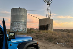In der Mitte des Bildes sieht man zwei runde Betontürme in der Landschaft, auf einem steht ein Windrad. Links im Bild sieht man die Motorhaube eines blauenn Jeeps. 