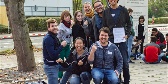 Eine Gruppe internationaler Studierender vor dem Internationalen Begegnungszentrum der TU Dortmund, die ein Spiel spielt; im Hintergrund eine weitere Gruppe