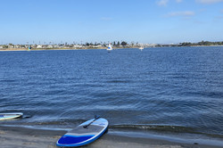 Ein blaues Stand-Up-Paddle-Board, das am Strand liegt. Im Hintergrund ist das Meer mit Häusern am Ufer und Segelbooten auf dem Wasser. 