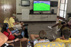 Hier sieht man einen Fernseher, auf dem Fußball läuft. Es sitzen mehrere Menschen auf Sofas um den Fernseher herum.