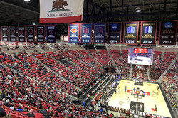 Ein Basketballstadion mit roter Tribüne rund um ein Basketballfeld. An der Decke hängen Fahnen und ein großer, runder Bildschirm, auf dem die Spieler zu sehen sind. 