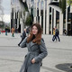 A portrait of a young woman. She has long brown hair, wears a gray coat and is standing in front of a building.  
