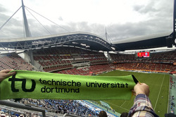 Hier sieht man den grünen TU-Schal, der in einem großen Fußballstadion hochgehalten wird. Im Hintergrund ist das Fußballfeld zu sehen. 