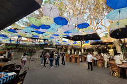 Hier sieht man einen großen Platz mit verschiedenen Restaurants. Auch hier hängen überall bunte Regenschirme.