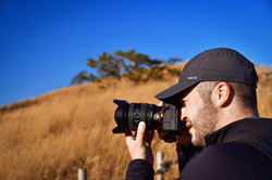 Storyteller Benedikt im Profil, wie wer mit einer Kamera ein Bild macht. Im Hintergrund sind gelb-braune Sträucher und blauer Himmel zu sehen. 