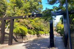 Hier sieht man einen asphaltierten Weg, rechts und links sind grüne Bäume und Büsche. Im Vordergrund ist ein Holztor auf dem japanische Schriftzeichen geschrieben stehen. 