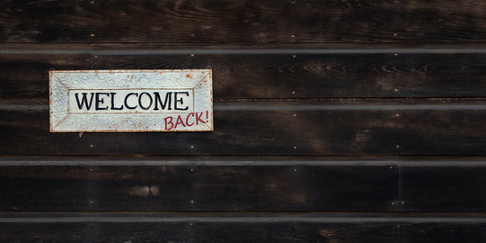 weißer "Welcome" Schild hängt am dunklen Holz. "Back" wurde handschriftlich hinzugefügt