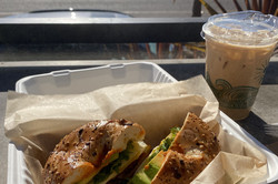 Zwei Bagel, belegt mit Avocado, Käse und Salat und ein Getränk auf einem kleinen Tablett. Im Hintergrund stehen Autos am Straßenrand und dahinter ist weite Landschaft. 