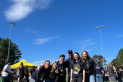 Eine Gruppe von Menschen posieren für die Kamera auf dem grünen Rasen eines Sportplatzes vor blauem Himmel. 