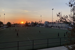 Hier sieht man einen großen Fußballplatz im Sonnenuntergang und Menschen, die auf dem Rasen Fußball spielen.