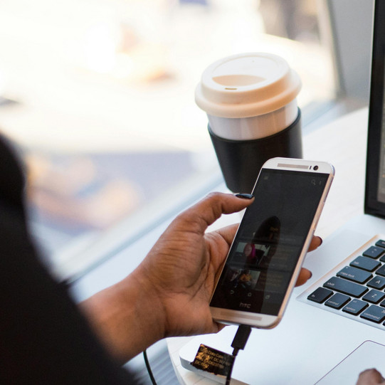 A woman is sitting at a round table in a café. On the table are a pair of glasses, a laptop, and a coffee mug. The woman is using her cell phone with her left hand and her laptop with her right hand.