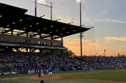 Hier sieht man ein Baseballspiel auf einem Platz in der freien Natur. Gerade geht die Sonne unter.