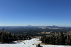 Hier sieht man eine Skipiste. die zwischen grünen Nadelbäumen den Berg hinabführt. Im Hintergrund sind kleine Berge zu sehen und der Himmel ist strahlend blau. 
