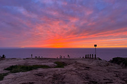 Gliders Point Man erkennt Klippen im Vordergrund und Meer im Hintergrund. Der Himmel ist orange gefärbt.