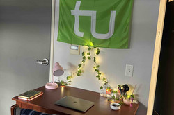 Schreibtisch im Dorm Das Bild zeigt einen Schreibtisch in einer Wohnung sowie eine Wand im Hintergrund, an der eine grüne Flagge der TU Dortmund hängt.