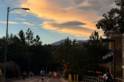 Eine Straße mit Studierenden im Vordergrund und den Bergen vor blauem Himmel, der durch den Sonnenuntergang orange gefärbten Wolken durchzogen ist.  
