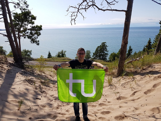 Daniel mit einer TU-Flagge vor einer Seelandschaft