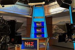 Hier sieht man ein Pult, das mit Scheinwerfern und Kameras umgeben ist. Im Hintergrund sind Bildschirme zu sehen, auf denen "NAZ Today" geschrieben steht. 