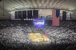 Hier sieht man ein gut gefülltes Stadion von Innen. Man erkennt den Basketballplatz in der Mitte und es hängt eine riesige USA Flagge von der Decke. 