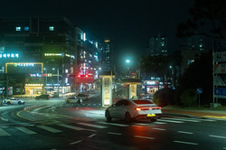 Große, vierspurige Straße bei Nacht, mit einem Taxi im Vordergrund und einer Straßenkreuzung sowie modernen Hochhäusern im Hintergrund. 