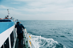 Ein Mann, der auf einem Boot vor Busan läuft. Ringsherum ist das Meer zu sehen und der Himmel ist leicht bewölkt. 