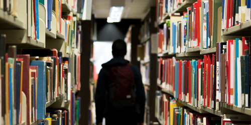 A male student stands in a library between two bookshelves