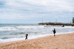 Ein Strand mit wilder Meeresbrandung. Eine Person fotografiert eine zweite Person, die vor den Wellen in Richtung Strand davonläuft. Im Hintergrund sind Brandungsmauern zu sehen. 