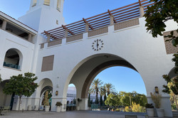 Conrad Prebys Aztec Student Union Hier sieht man einen großen Rundbogen mit einer Uhr über einem gepflasterten Platz. Links am Rundbogen steht ein großer, weißer Turm im spanischen Baustil.