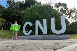 Storyteller Benedikt hält eine grüne TU-Flagge in der Hand und steht neben den großen, weißen Buchstaben "CNU" vor grünen Bäumen im Hintergrund. 