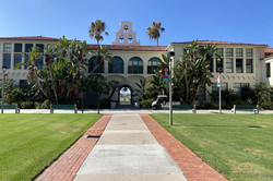 Hepner Hall In der Mitte ist ein großes Gebäude mit vier Rundbögen über einem Eingangstor. Dorthin führt ein gepflasterter Weg, umgeben von grüner Wiese und zwei großen Palmen.