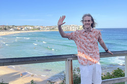 Bondi Beach Will winkt in die Kamera und steht an einem Aussichtspunkt hoch über einem Strand
