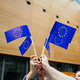 Menschen stehen in einem Kreis und halten kleine EU-Flaggen in der Luft.