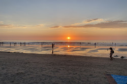 Ein Strand im Sonnenuntergang mit einigen Menschen, die im Meer stehen oder am Strand spazieren gehen. 
