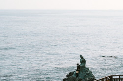 Ausblick auf das Meer bei diesiger Sicht. Im Vordergrund ist eine Meerjungsfrauenstatue auf einem Felsvorsprung zu sehen. 