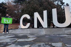 Hier sieht man Benedikt neben den drei Buchstaben "CNU". Er hält eine grüne Flagge mit dem TU-Logo in der Hand. 