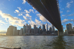 NYC Hier sieht man zahlreiche Hochhäuser bei blauem Himmel. Man sieht Wasser und eine Brücke von unten.