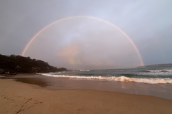 Es zeigt einen großen Regenbogen an einem Sandstrand, das Meer schlägt Wellen.
