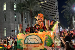 Hier sieht man einen Umzugswagen für Halloween bei Nacht. Präsent sind ein Totenkopf auf dem Wagen und mehrere verkleidete Menschen. 