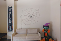 Hier sieht man ein Zimmer mit Sofa und darüber ist ein Spinnennetz als Deko, genauso wie eine aufblasbare Hexe und mehrere Kürbisse.