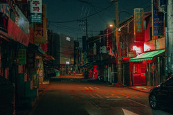 Eine schmale Straße bei Nacht mit vielen bunten Schildern mit koreanischen Schriftzeichen rechts und links an den Gebäuden und Stromleitungen über der Straße. 