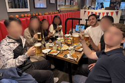Hier sieht man eine Gruppe von Menschen, die um einen Tisch herumsitzen und mit ihren Getränken in Richtung Kamera "prosten".