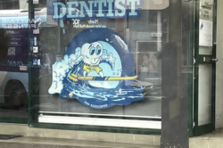 Es zeigt ein Schaufenster mit einem Zahnarztlogo: Ein Zahn mit Gesicht surft auf einer Zahnbürste, oben steht die Überschrift Dentist